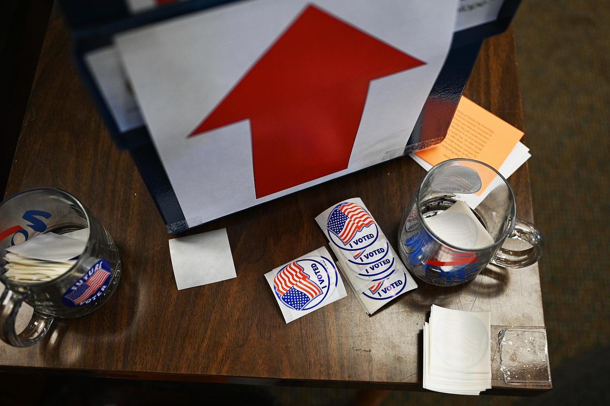 Table with "I Voted" stickers, mugs, and a large red arrow sign at a voting location.